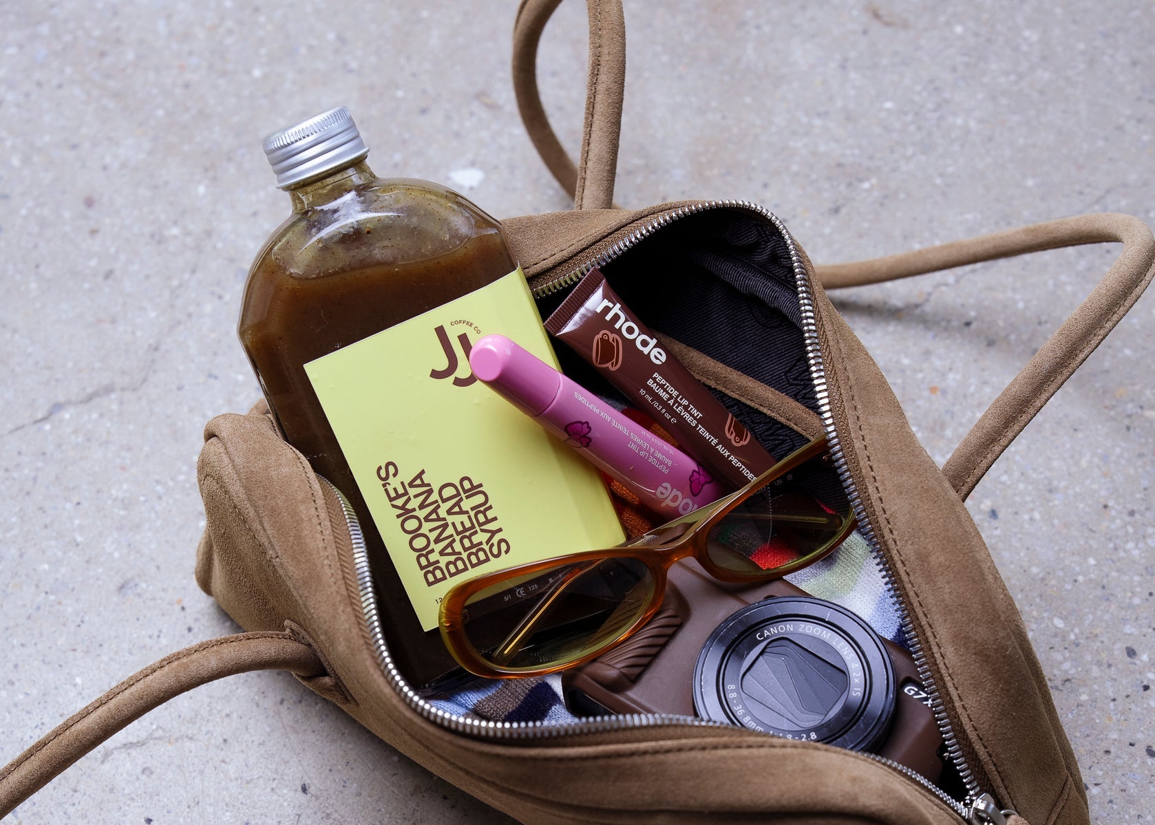 Open beige handbag with various items including a syrup bottle, sunglasses, and a ribbon on a concrete surface.