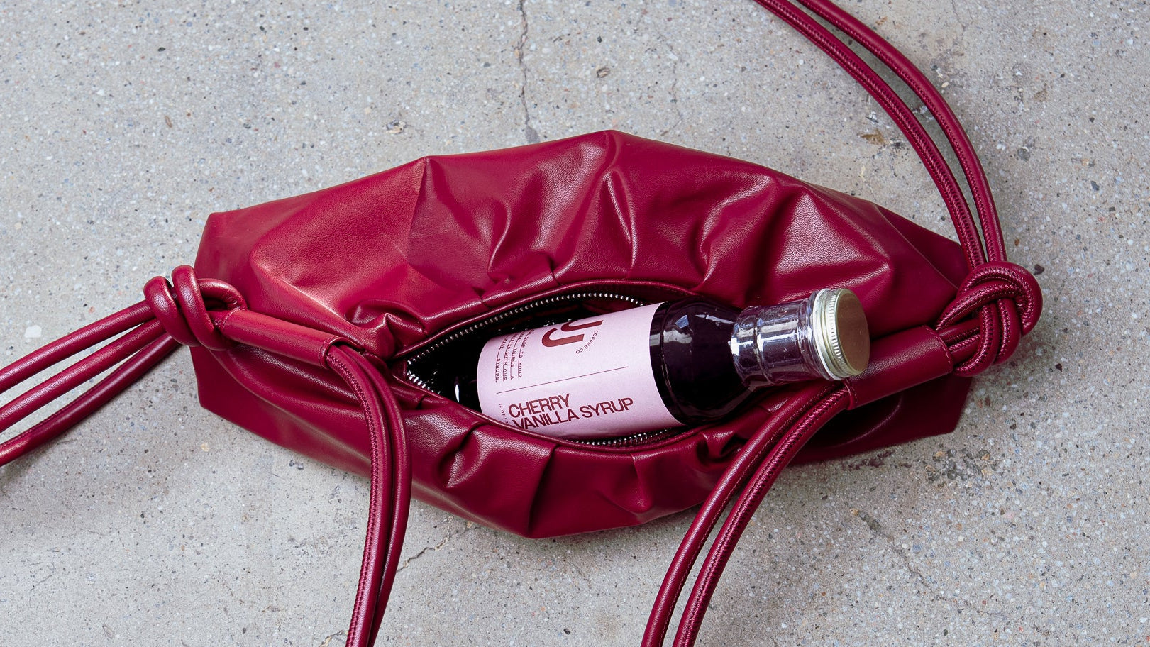 Red leather bag with a cherry vanilla syrup bottle inside on a concrete surface